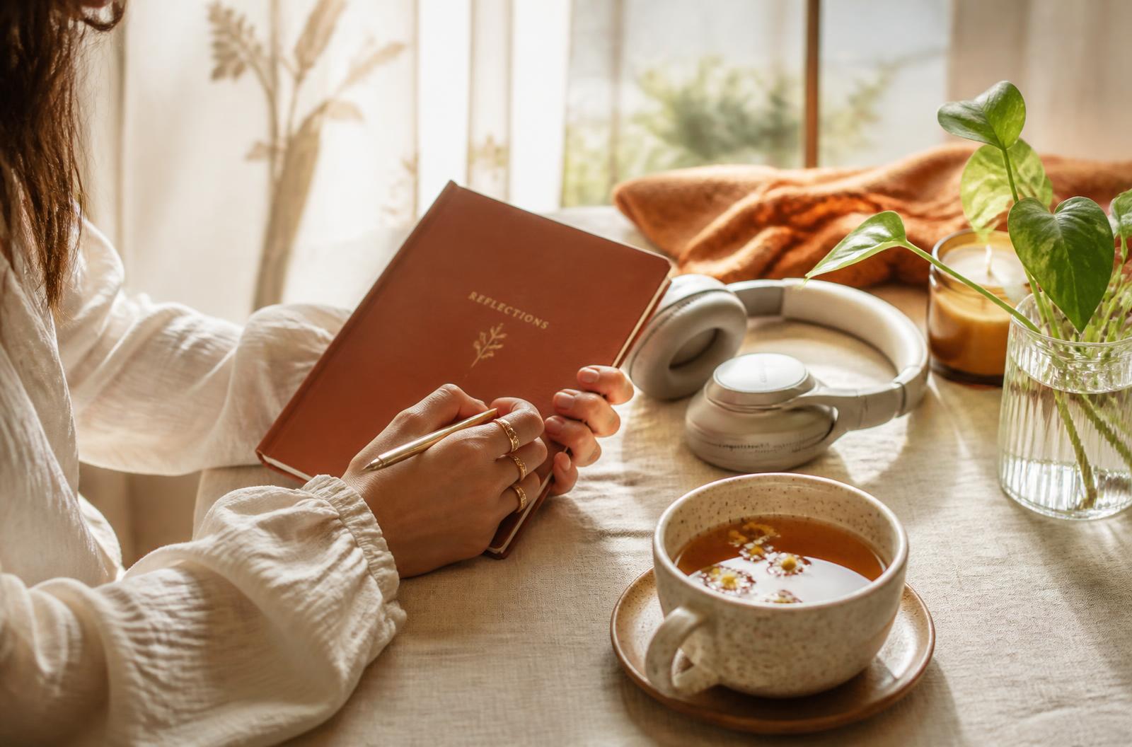 Manos de mujer escribiendo en un cuaderno junto a una taza de té y unos auriculares